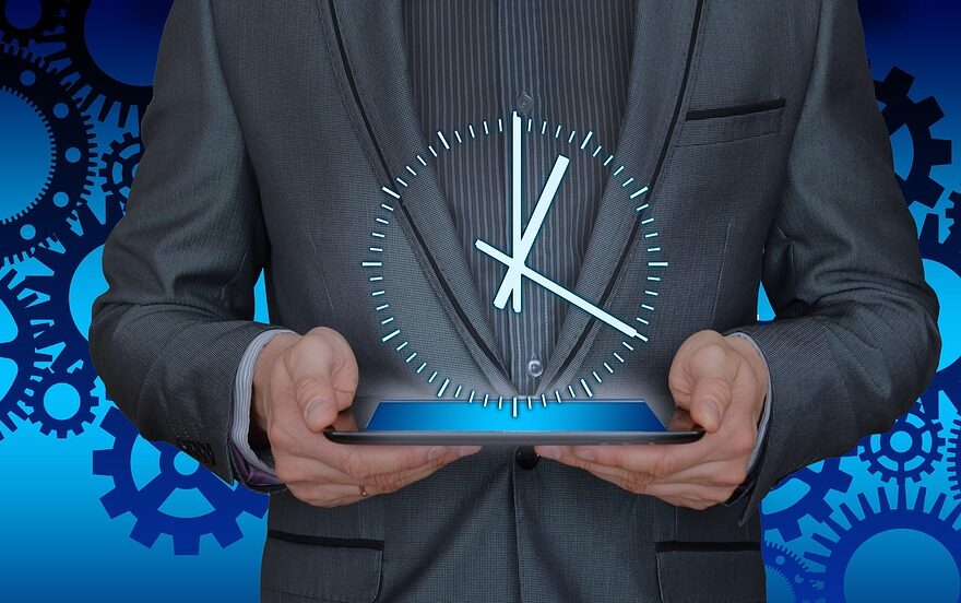 Close-up shot of a man in a gray shirt jacket holding a tablet projecting a clock illustration.