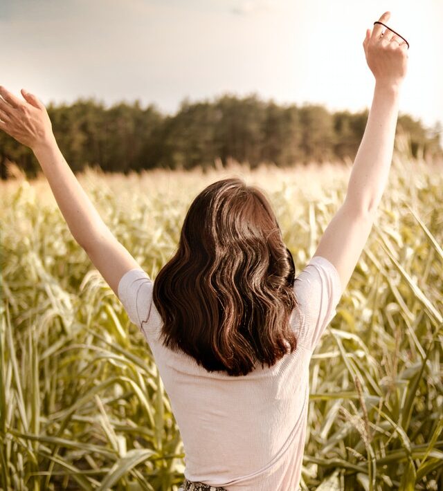 Back view of a woman with curly brown hair and arms in the air in a wheat field.