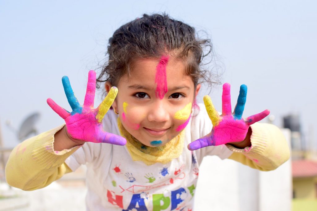 Portrait of a young girl covered in colored paint with her hands open and stains on both sides of her face.